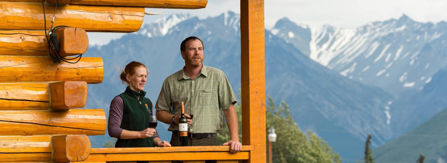 A couple enjoys a scenic view of snow-capped mountains from a rustic wooden deck in Mat-Su, Alaska