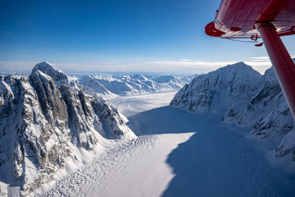 A breathtaking aerial view of a vast glacier surrounded by towering snow-capped mountains under a clear blue sky in Mat-Su, Alaska