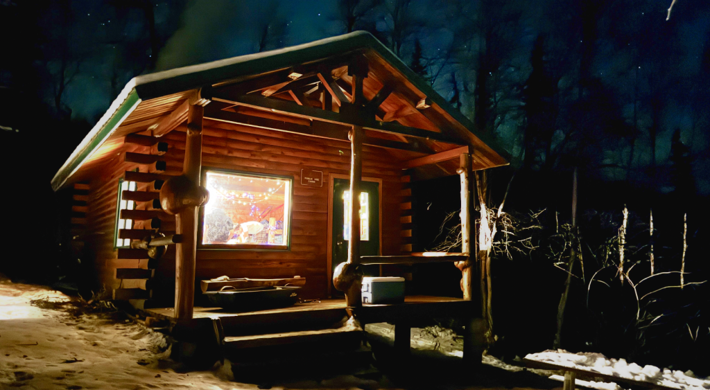 A cozy log cabin illuminated at night in a snowy forest setting in Mat-Su, Alaska