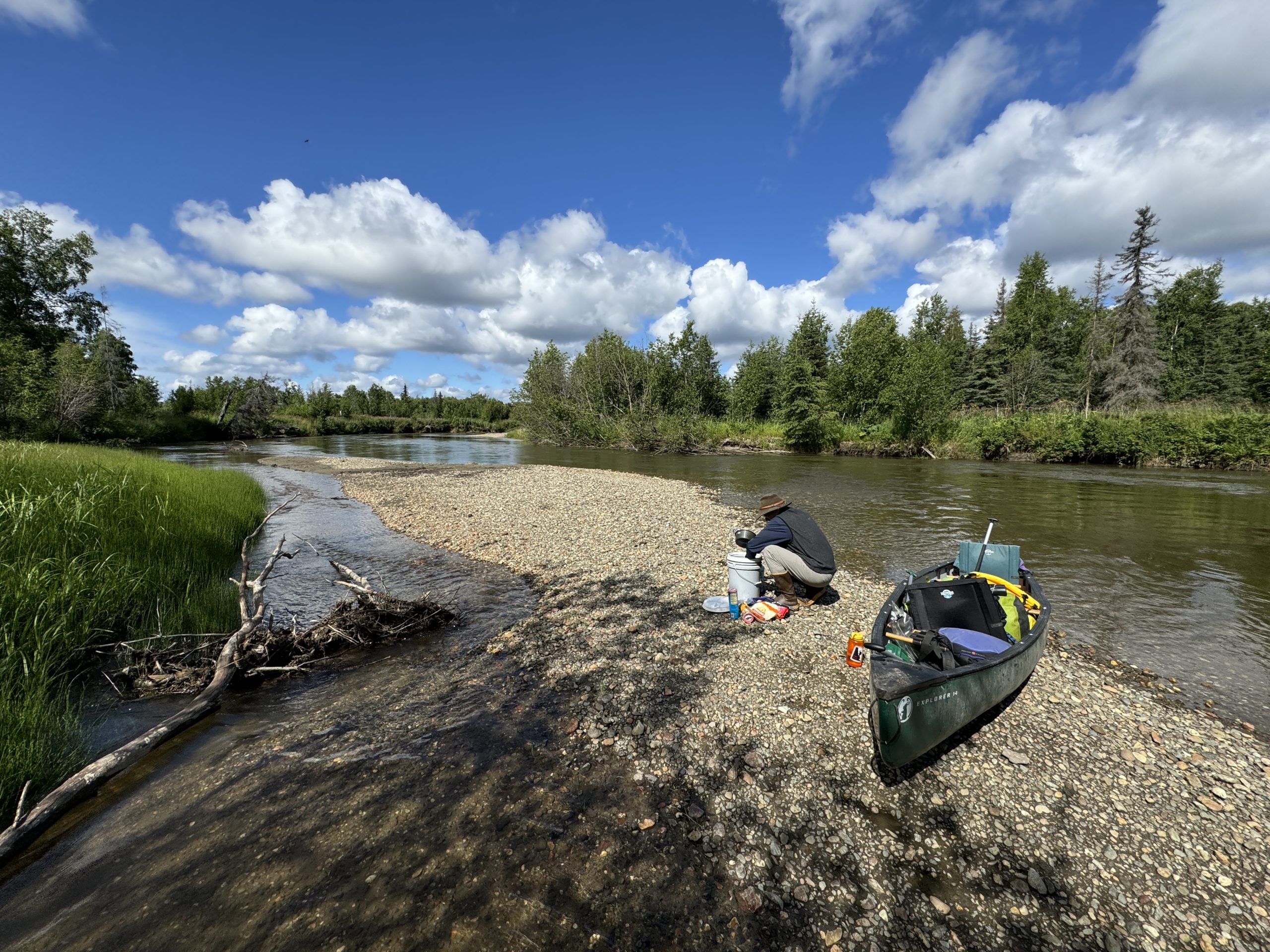 A person rests by their canoe on a gravel riverbank under a cloudy sky in Mat-Su, Alaska