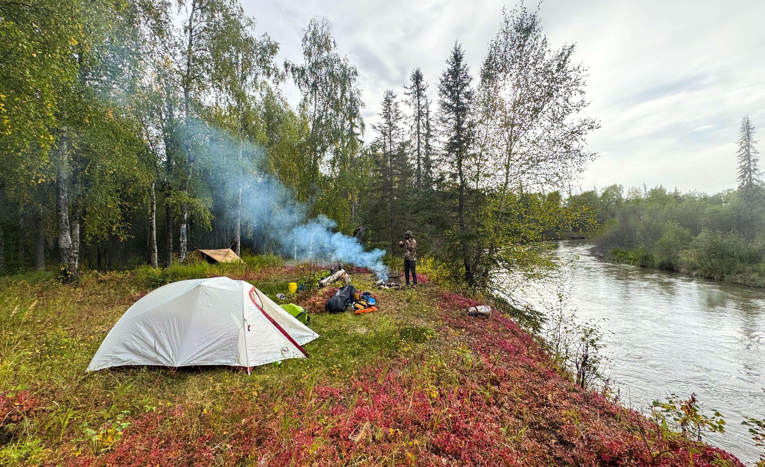 A white tent set up by a river with smoke rising from a campfire in Mat-Su, Alaska