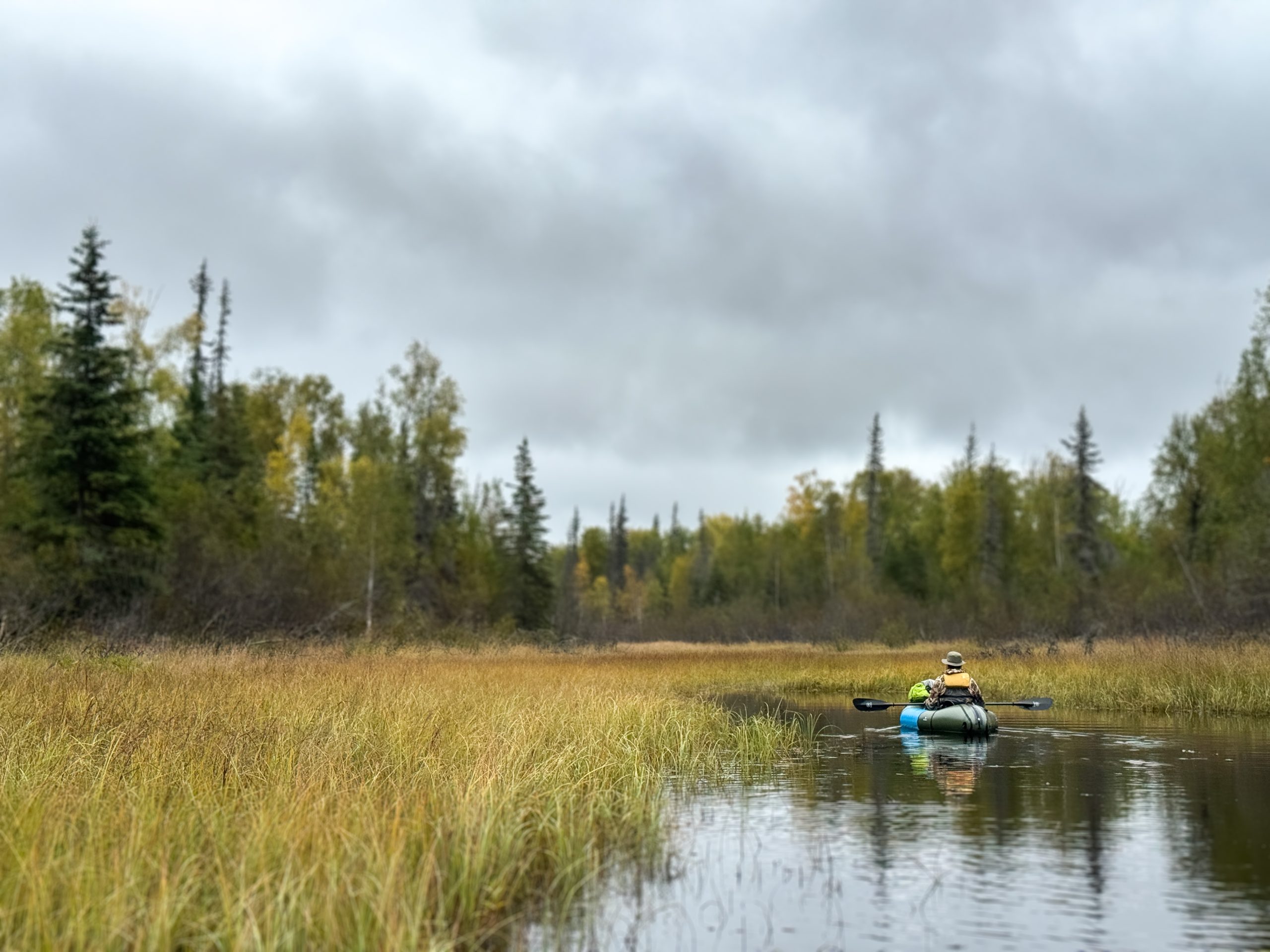 A person paddles a small raft through a calm, grassy waterway under cloudy skies in Mat-Su, Alaska