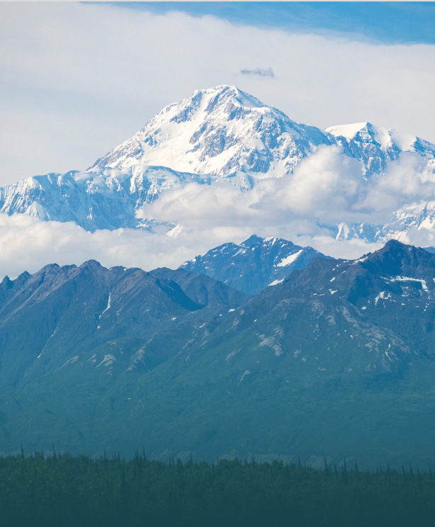 A majestic snow-capped mountain peak rises above a range of rugged, forested mountains in Mat-Su, Alaska
