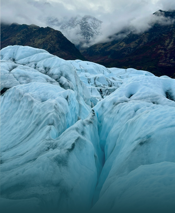 A vast glacier with deep blue crevasses stretches across a rugged landscape, framed by distant snow-capped mountains in Mat-Su, Alaska