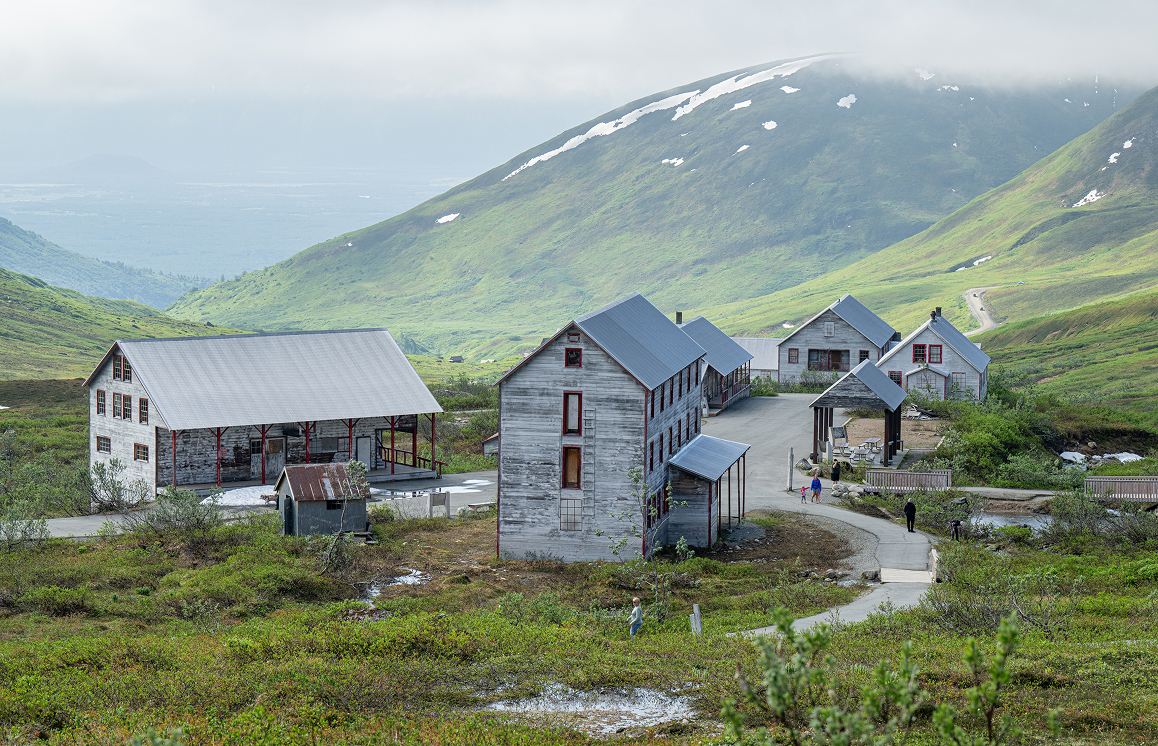 A cluster of rustic buildings nestled in a lush green valley with snow-capped mountains in the distance in Mat-Su, Alaska