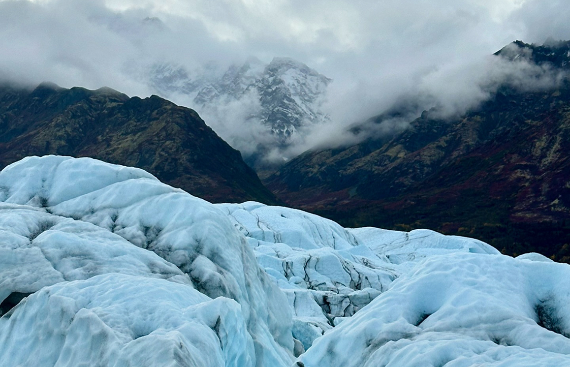 A vast expanse of jagged, blue ice stretches across a mountainous landscape, with misty peaks rising in the distance in Mat-Su, Alaska