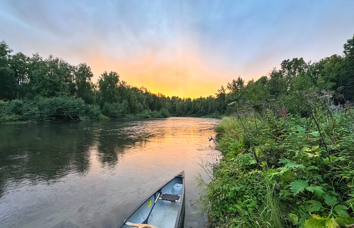 A serene river scene at sunset with a canoe resting on the shore, surrounded by lush greenery and a tranquil sky in Mat-Su, Alaska