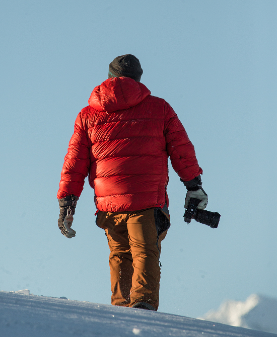 A person in a bright red puffer jacket walks across a snowy landscape, carrying a camera in Mat-Su, Alaska