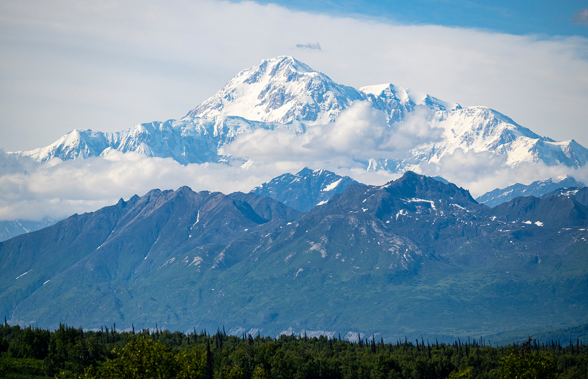 A majestic snow-capped mountain peak rises above a lush green forest, with clouds swirling around its base in Mat-Su, Alaska
