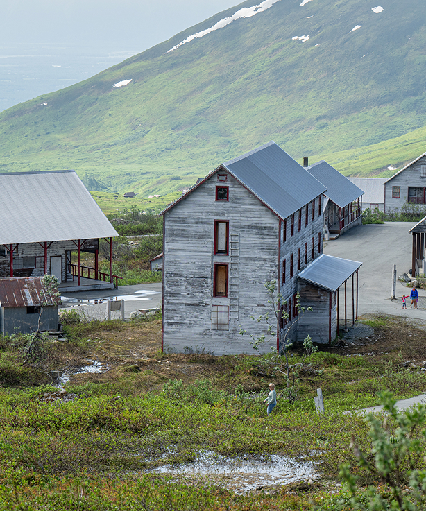 A rustic wooden building with a blue roof stands amidst lush green hills and a misty mountain backdrop in Mat-Su, Alaska