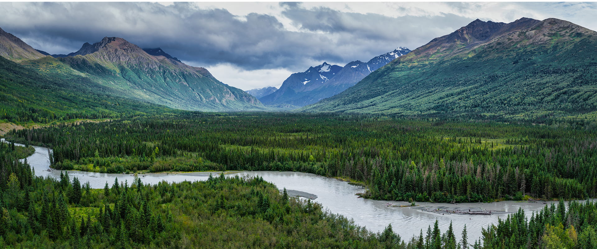 A serene landscape featuring a winding river surrounded by lush green forests and towering mountains with snow-capped peaks under a cloudy sky in Mat-Su, Alaska