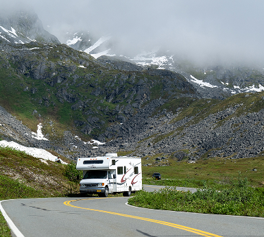 A white RV travels a winding mountain road with snow-capped peaks in the background in Mat-Su, Alaska