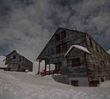 Two weathered wooden buildings stand in a snowy landscape under a cloudy sky in Mat-Su, Alaska