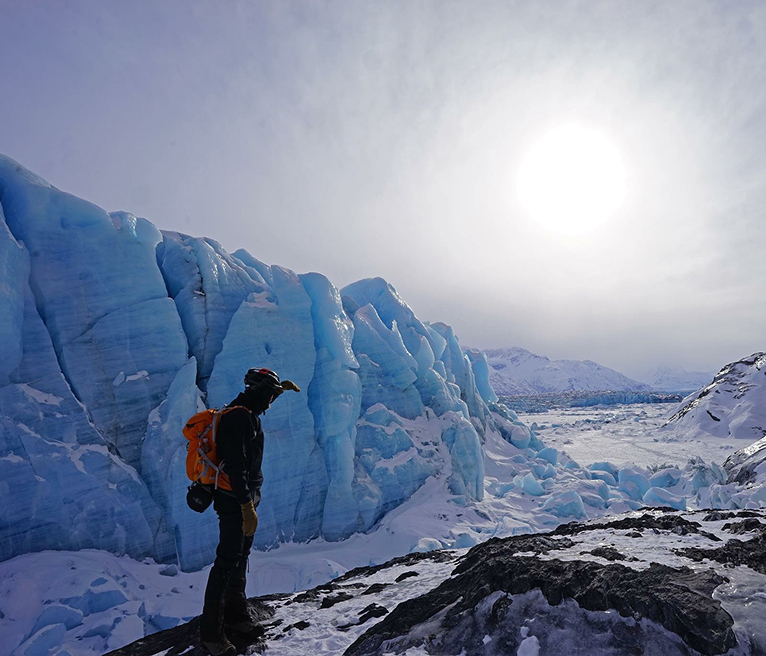 A lone hiker stands on a rocky outcrop, gazing at towering blue ice cliffs under a bright sun in Mat-Su, Alaska