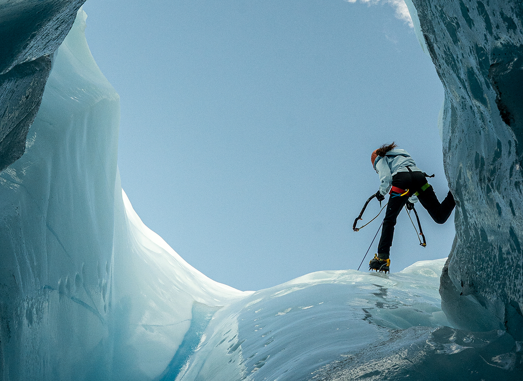 A climber ascends a steep ice wall, surrounded by towering ice formations, under a clear blue sky in Mat-Su, Alaska