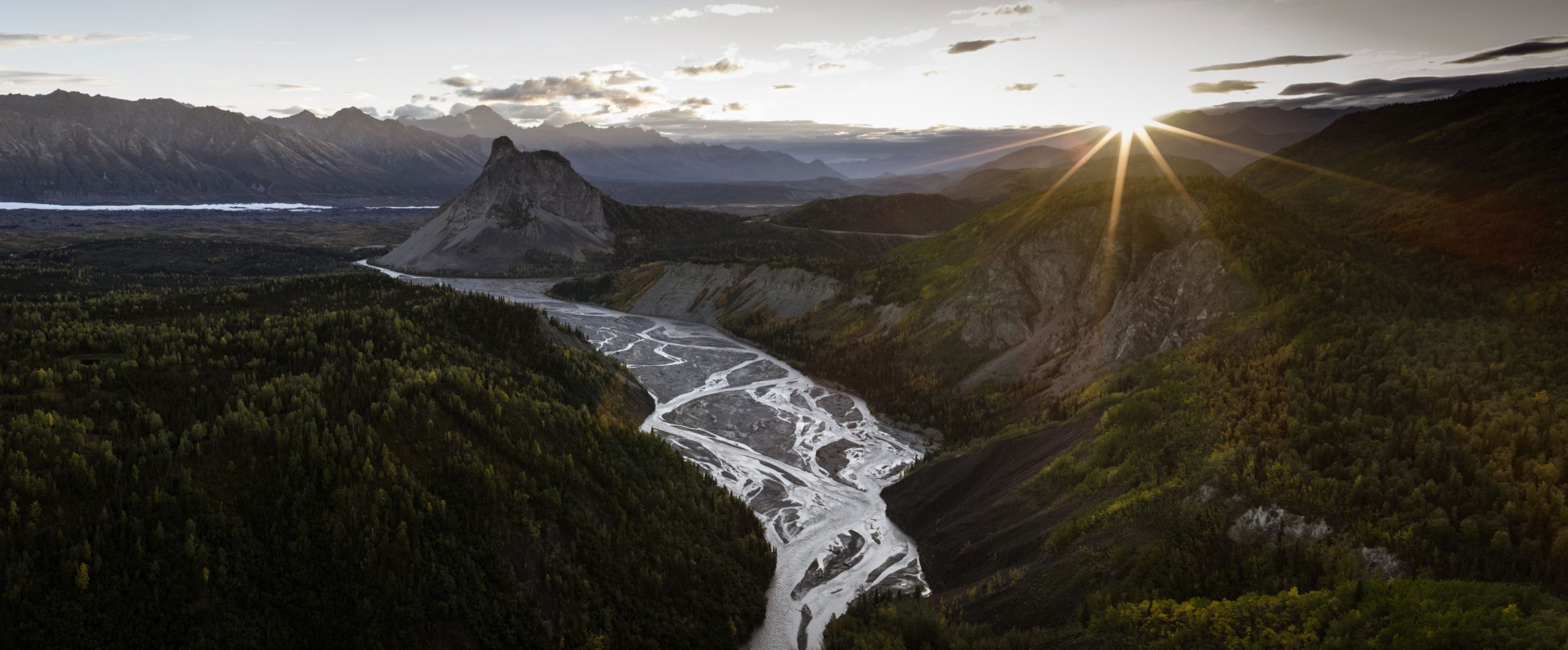 A panoramic view of a winding river cutting through a lush, green valley with majestic mountains in the background during sunset in Mat-Su, Alaska