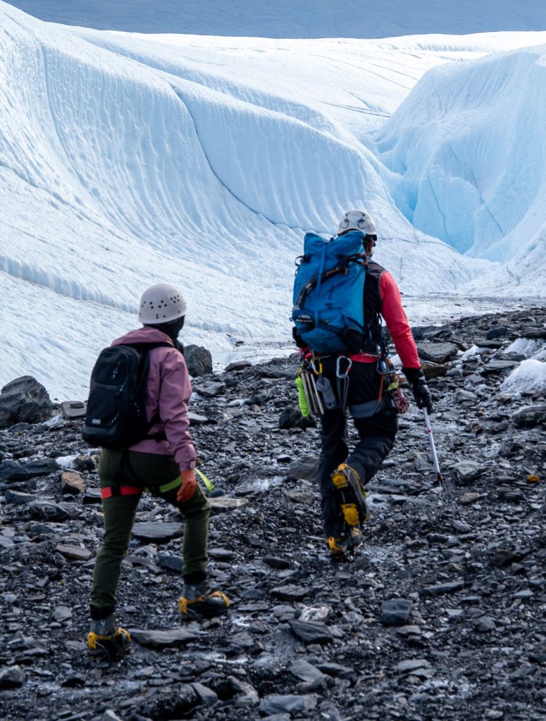 Two hikers, equipped with backpacks and ice axes, navigate a rocky glacier with towering ice formations in the background in Mat-Su, Alaska