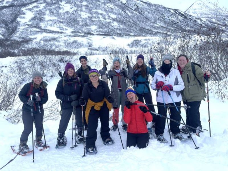 A group of ten people, dressed in winter clothing, standing in a snowy landscape with snowshoes and ski poles, smiling at the camera in Mat-Su, Alaska