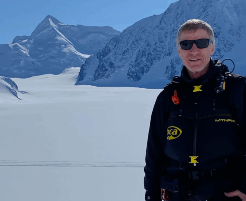 A man stands in front of a breathtaking snow-capped mountain range under a clear blue sky in Mat-Su, Alaska