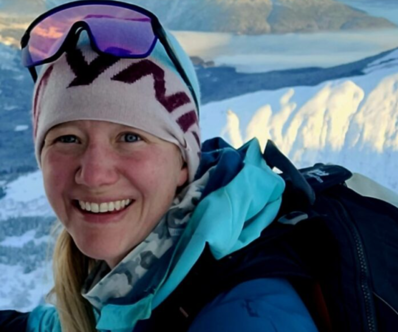 A smiling woman wearing a beanie, sunglasses, and a blue jacket stands amidst a snowy landscape with mountains in the background in Mat-Su, Alaska