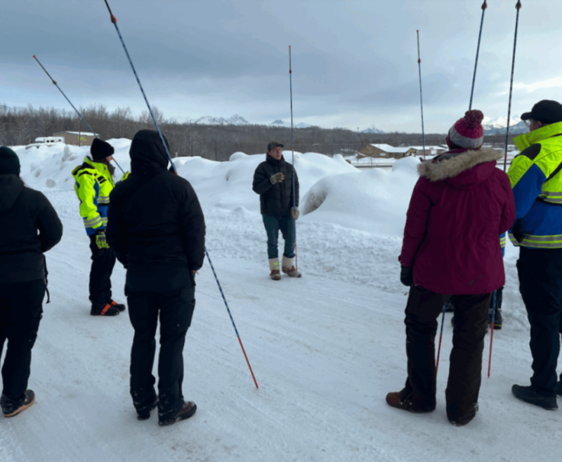 A group of people dressed in winter gear are gathered in a snowy landscape, listening to a speaker who is standing in front of them in Mat-Su, Alaska