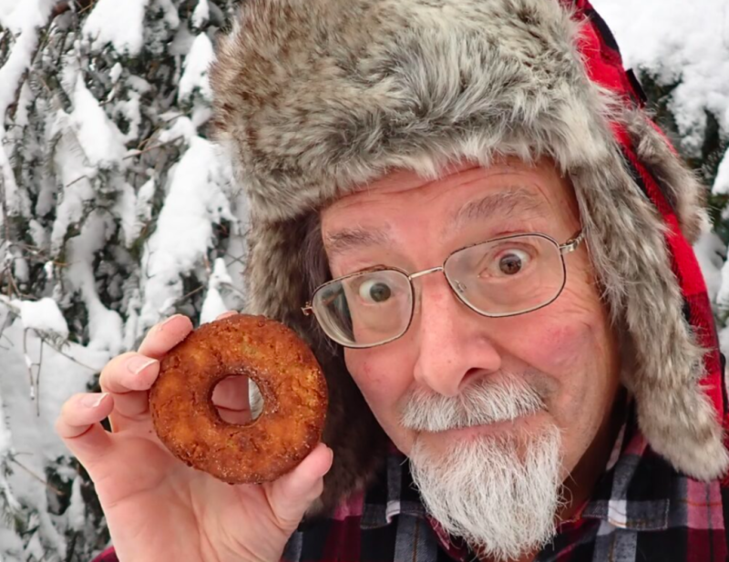 An elderly man with a white beard and glasses, wearing a fur-lined hat, holds up a donut in a snowy forest setting in Mat-Su, Alaska