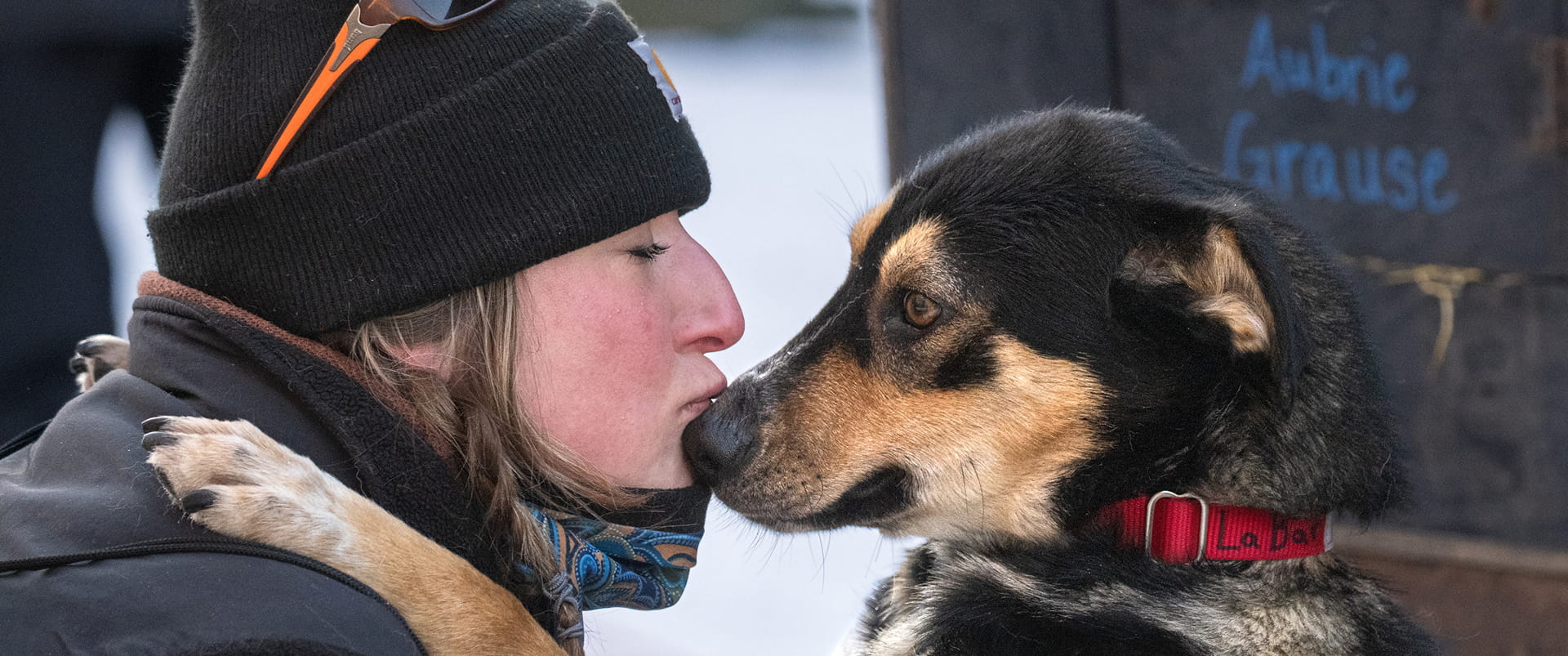 A woman wearing a black beanie and sunglasses embraces a black and tan dog with a red collar, sharing a tender moment in a snowy setting in Mat-Su, Alaska