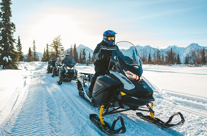 A group of snowmobilers riding through a snowy forest with mountains in the background in Mat-Su, Alaska