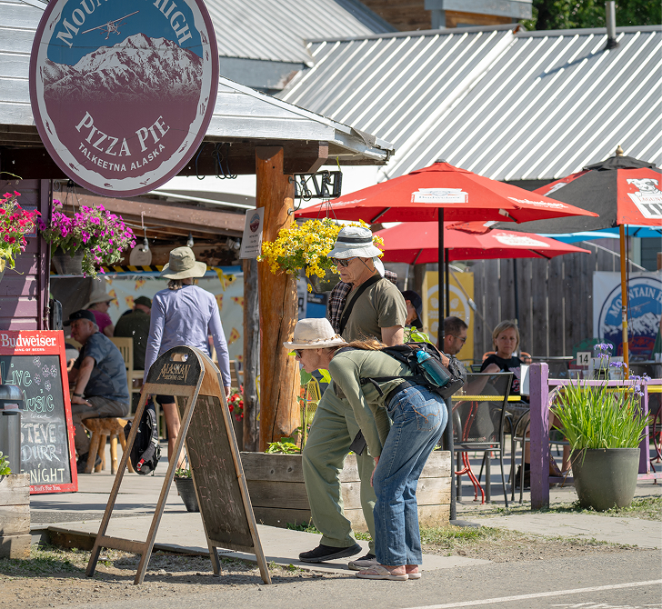 A lively outdoor cafe with colorful umbrellas, potted plants, and a chalkboard menu, set against a backdrop of mountains in Mat-Su, Alaska