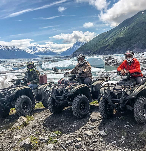 Three ATV riders in full gear explore a rugged, icy landscape with snow-capped mountains in the distance in Mat-Su, Alaska