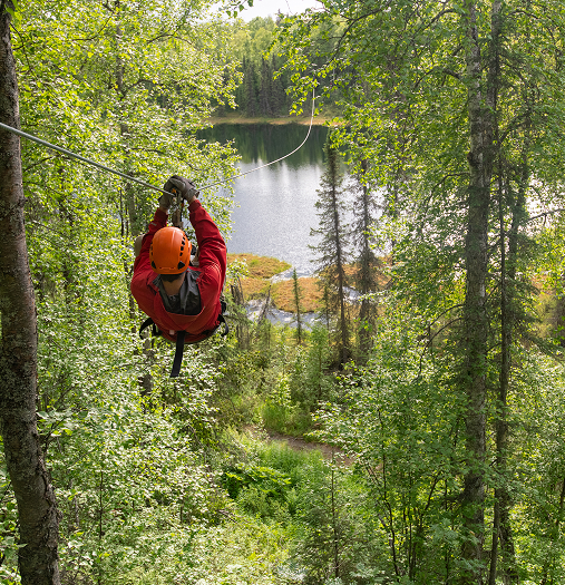 A person in a red helmet zips through the air on a zipline over a serene lake surrounded by lush green forest in Mat-Su, Alaska