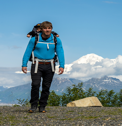 A hiker with a baby in a carrier on his back walks along a gravel path with snow-capped mountains in the distance in Mat-Su, Alaska