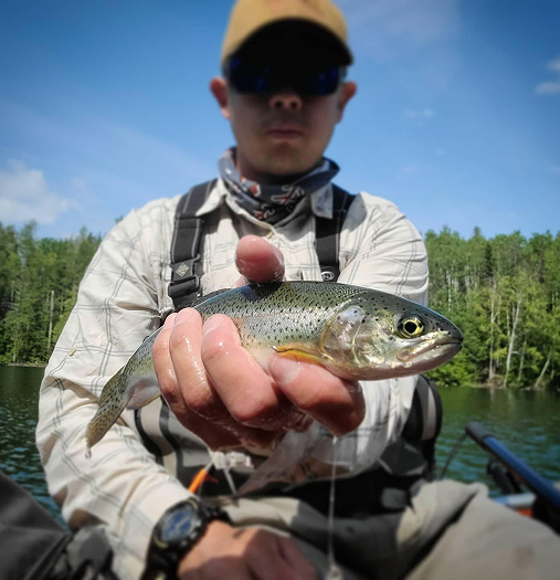 A man proudly holds up a freshly caught fish on a serene lake surrounded by lush greenery in Mat-Su, Alaska