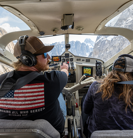 Two pilots in a helicopter cockpit with snowy mountain peaks visible through the windshield in Mat-Su, Alaska