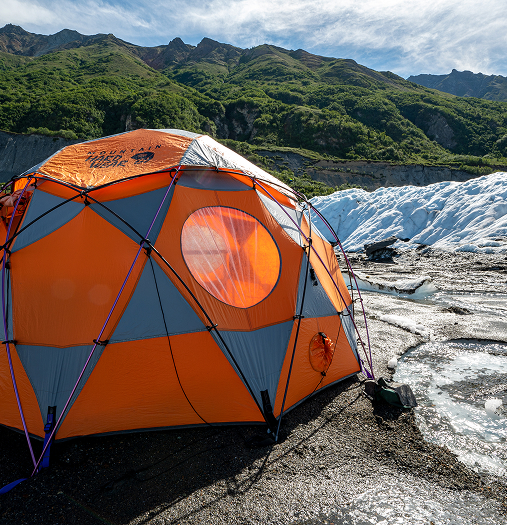 An orange and gray dome tent set up on a rocky, icy terrain with a backdrop of snow-capped mountains and lush green hills in Mat-Su, Alaska