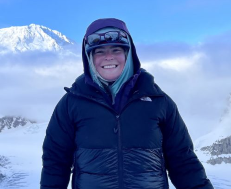 A person smiling in front of a snow-capped mountain, wearing a black jacket, hood, and sunglasses in Mat-Su, Alaska