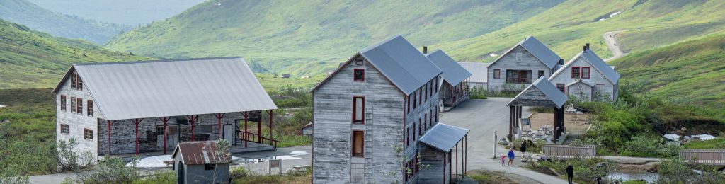 A cluster of rustic wooden buildings nestled in a lush green valley surrounded by rolling hills in Mat-Su, Alaska