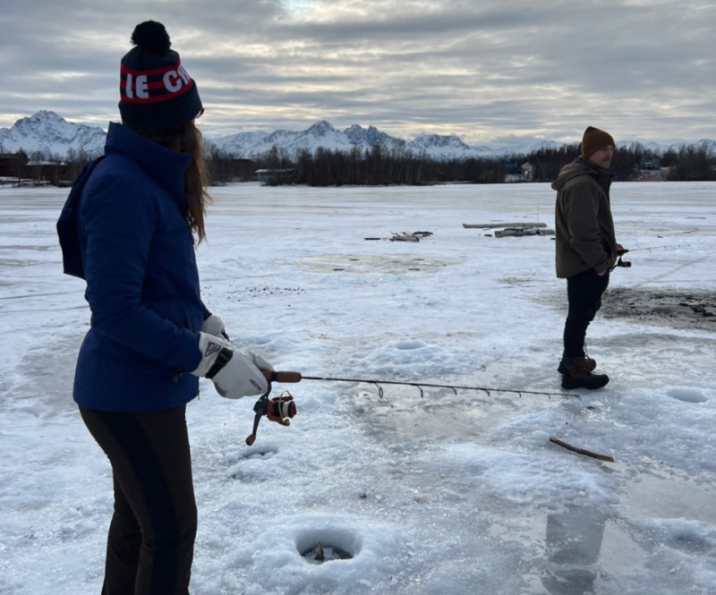 Two people ice fishing on a frozen lake with mountains in the background in Mat-Su, Alaska