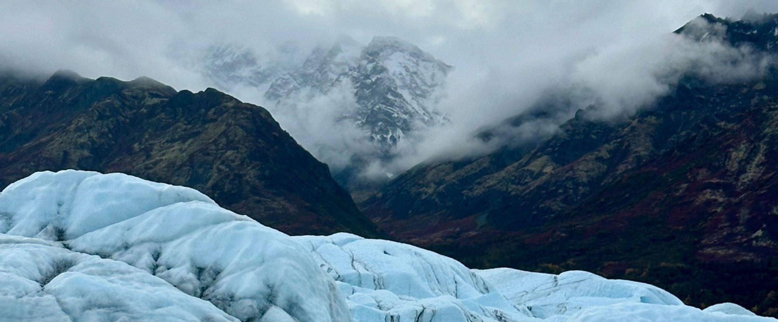 A serene mountain landscape with snow-capped peaks shrouded in mist, and a foreground of jagged ice formations in Mat-Su, Alaska