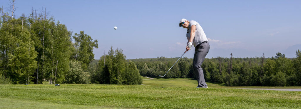 A golfer in mid-swing on a lush green course, with a ball in the air and a scenic backdrop of trees and blue sky in Mat-Su, Alaska