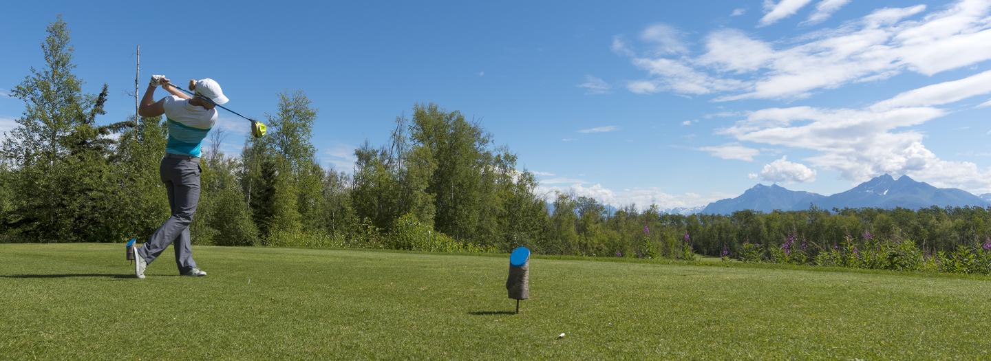 A golfer in mid-swing on a lush green course with a scenic mountain backdrop in Mat-Su, Alaska