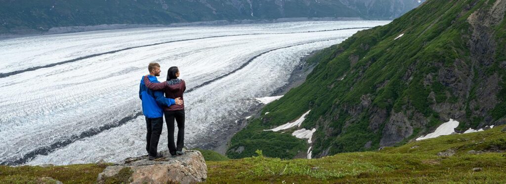 A couple stands on a rocky outcrop overlooking a vast glacier with snow-capped mountains in the distance in Mat-Su, Alaska