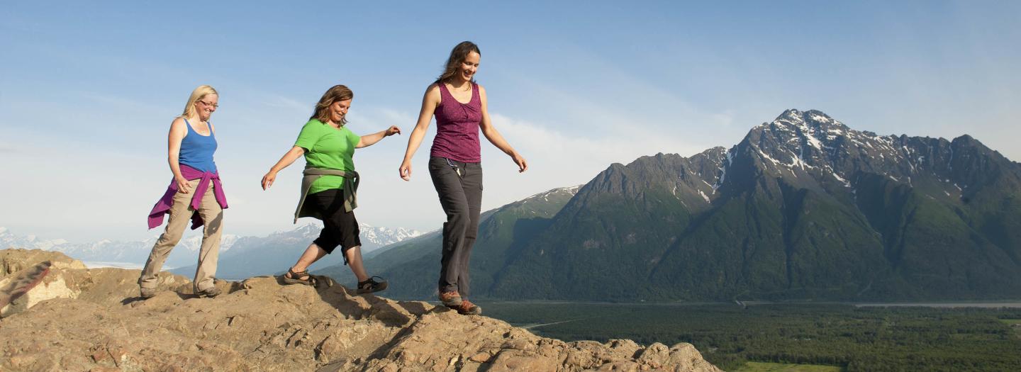 Three women hike along a rocky mountain ridge with snow-capped peaks in the distance in Mat-Su, Alaska