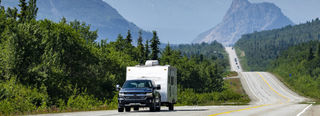A pickup truck towing a white camper travels down a winding mountain road surrounded by lush green forests and towering peaks in Mat-Su, Alaska