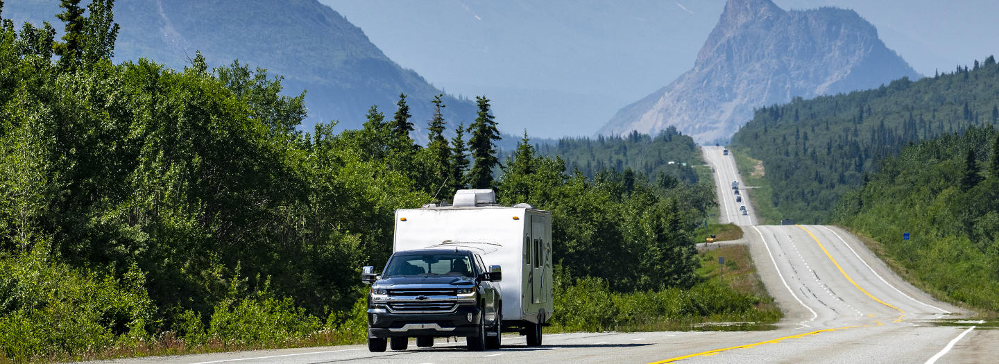 A pickup truck towing a white camper travels down a winding mountain road surrounded by lush green forests and towering peaks in Mat-Su, Alaska