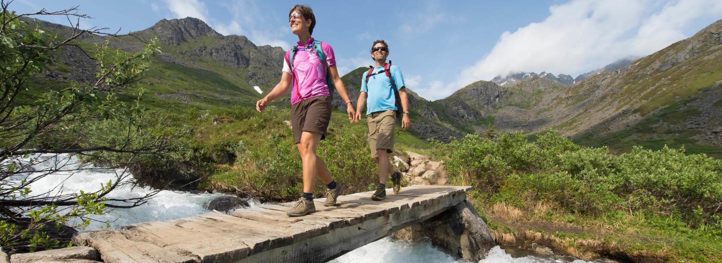 Two hikers cross a wooden bridge over a rushing mountain stream, surrounded by lush green hills and snow-capped peaks in Mat-Su, Alaska