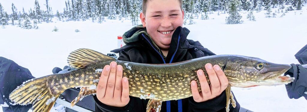 A young boy proudly displays a large walleye fish caught in a snowy, winter landscape in Mat-Su, Alaska