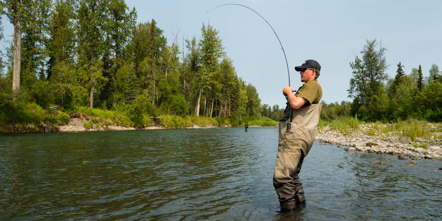 A man in waders stands in a river, casting a fishing rod amidst a serene, tree-lined landscape in Mat-Su, Alaska