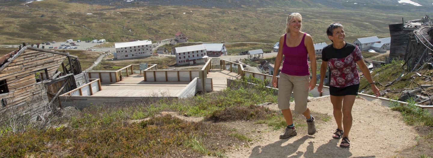 Two women walking along a dirt path in a mountainous area with abandoned buildings in the background in Mat-Su, Alaska