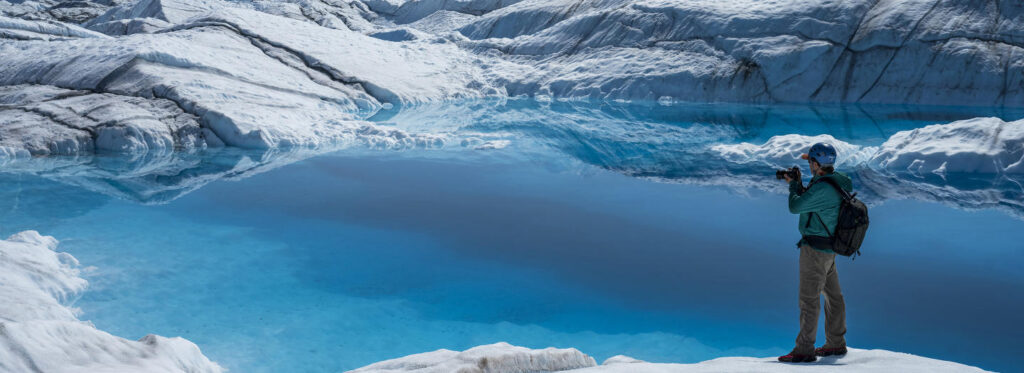 A lone hiker captures a breathtaking view of a serene, turquoise glacial lake surrounded by snow-capped mountains in Mat-Su, Alaska